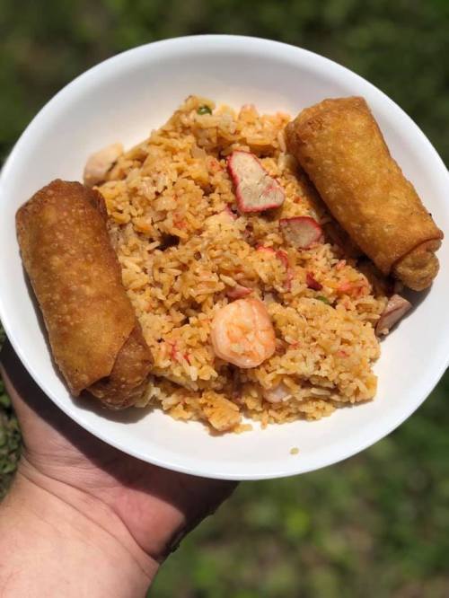 A hand holding a bowl of fried rice with shrimp and two crispy spring rolls on the side.