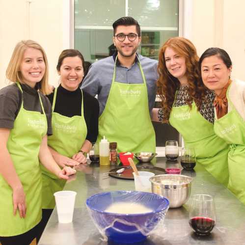 A group of five people in green aprons smiles around a kitchen counter with cooking ingredients and utensils.