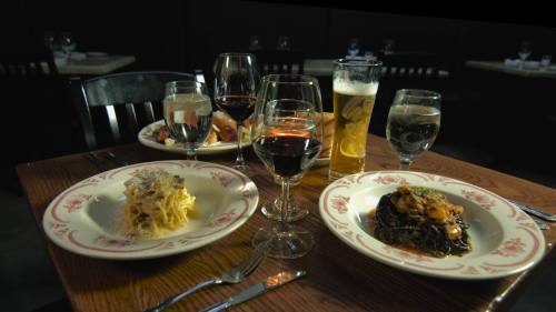 A table set with two plates of pasta, glasses of wine, water, and a beer, in a cozy restaurant setting.