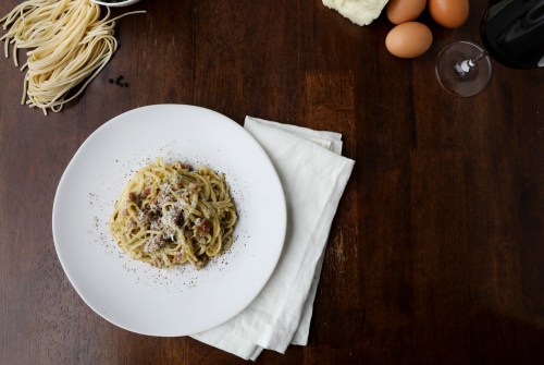 A plate of pasta with cheese and pepper, surrounded by ingredients like eggs, wine, and uncooked pasta on a wooden table.
