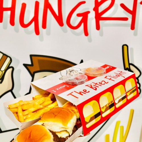 A box labeled "The Bitez Flight" containing sliders and fries, with dipping sauces, against a colorful wall.