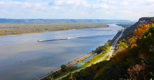 A scenic view of a river winding through green hills, with a barge traveling along the water and a road beside it.
