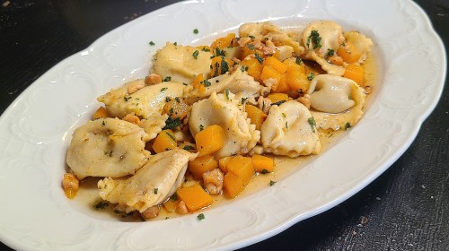 A white platter filled with ravioli, butternut squash, and chopped nuts, garnished with herbs in a light sauce.