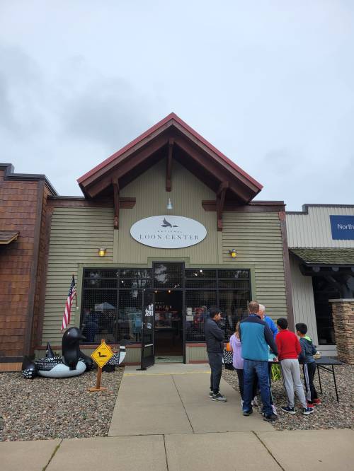 A group of people stands outside the Loon Center, a wooden building with a sign and an American flag.
