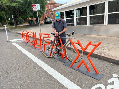 A person adjusts their bike next to a colorful bike rack with the word "COLE" in bold letters on a city street.