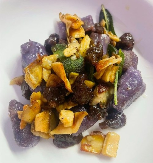 A plate of purple gnocchi topped with sautéed apples, nuts, and herbs.