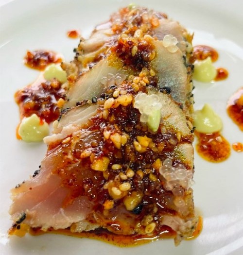 Sliced fish topped with a spicy sauce, sesame seeds, and dollops of creamy sauce on a white plate.
