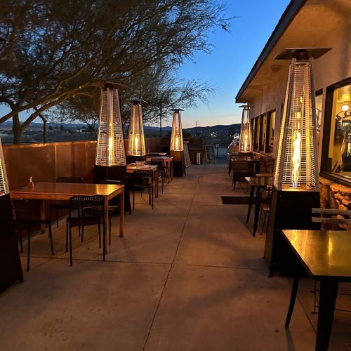 Outdoor dining area with tall heaters, tables, and chairs, surrounded by trees and mountains under a clear sky.