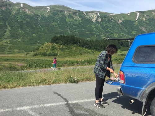 A person prepares food by a blue truck in a grassy area, with a mountain backdrop and another person walking in the distance.