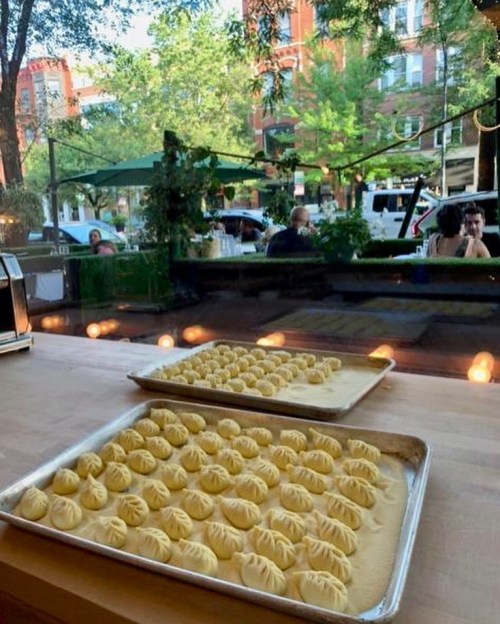 Trays of dumplings on a wooden table, with a view of a lively outdoor dining area and greenery in the background.