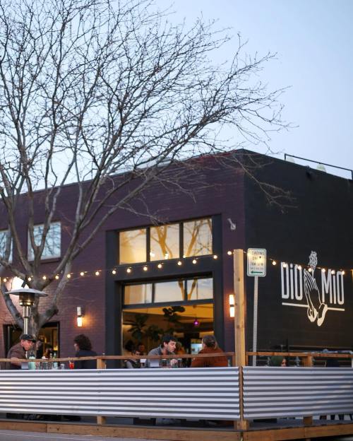 Outdoor dining area of a restaurant with string lights, a tree, and a modern building facade at dusk.