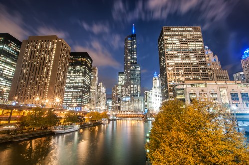 City skyline at night with illuminated skyscrapers reflecting on a river, surrounded by autumn trees.