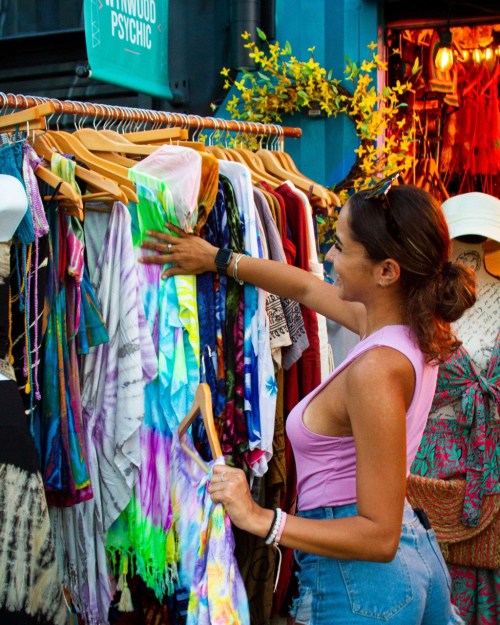 A woman browses colorful tie-dye clothing on display at a market, surrounded by vibrant decorations.