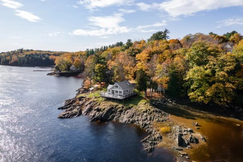 A scenic view of a house on a rocky shore, surrounded by vibrant autumn foliage and a calm river.
