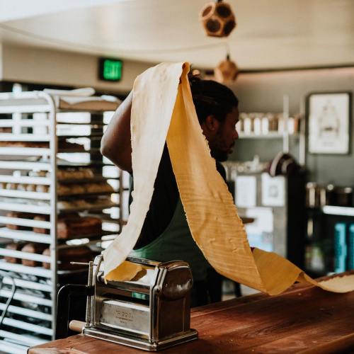 A chef stretches a long sheet of pasta dough in a kitchen, with baking racks in the background.