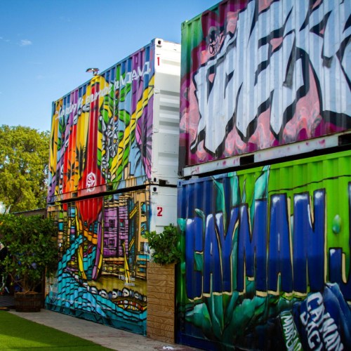 Colorful graffiti on shipping containers, surrounded by greenery and a blue sky.
