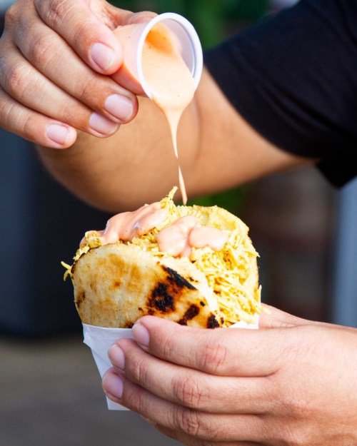 A hand holds a taco filled with shredded meat, while drizzling sauce from a small cup on top.