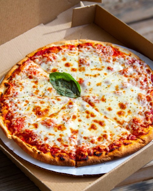 A freshly baked cheese pizza with a single basil leaf, cut into slices, in a cardboard pizza box.