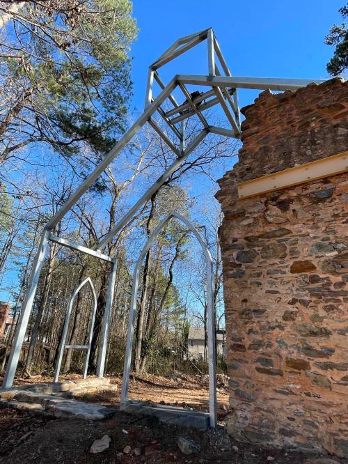 A partially collapsed stone structure with a metal frame resembling a church, surrounded by trees and blue sky.