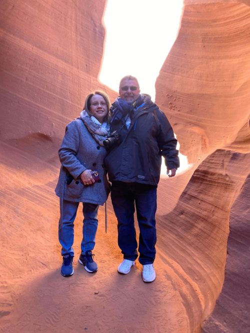 A couple stands together in a narrow, sunlit canyon, surrounded by smooth, reddish rock formations.