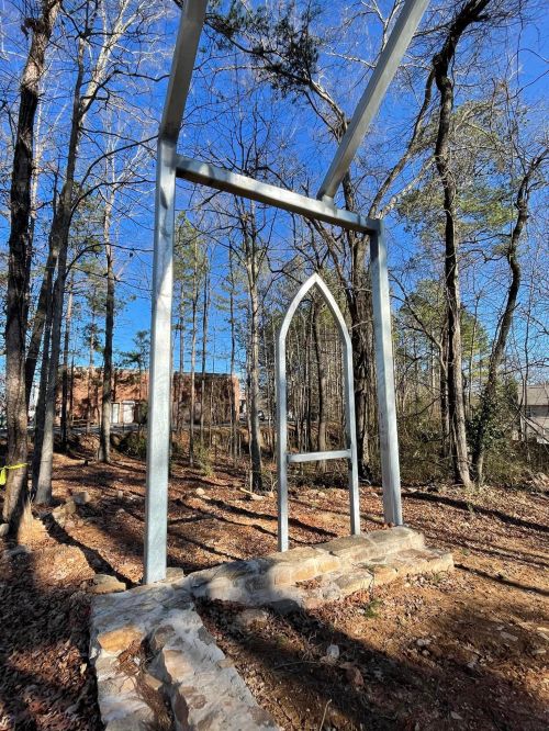 A metal frame resembling a gothic arch stands in a wooded area, surrounded by trees and fallen leaves.