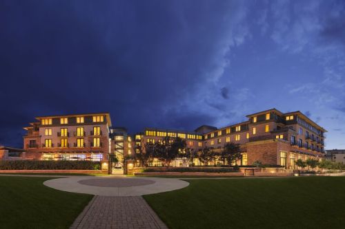 A modern hotel building illuminated at dusk, surrounded by green lawns and a circular pathway.