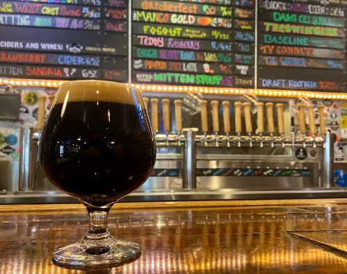 A dark beer in a glass sits on a wooden bar, with a colorful beer menu and taps in the background.