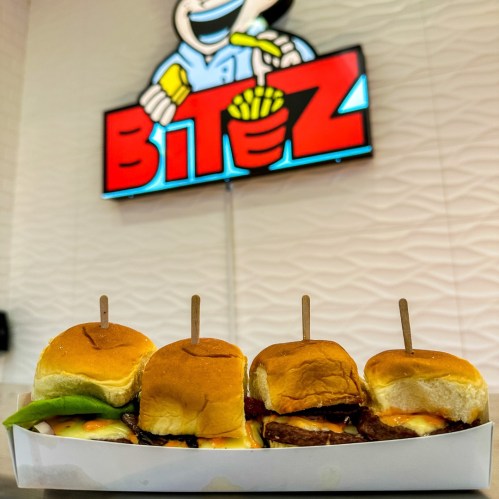 Four sliders on a tray with a colorful "BITEZ" sign in the background, featuring a cartoon character holding fries.