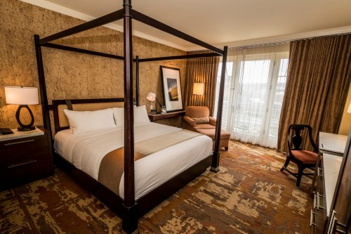 A cozy hotel room featuring a four-poster bed, armchair, desk, and large windows with natural light.