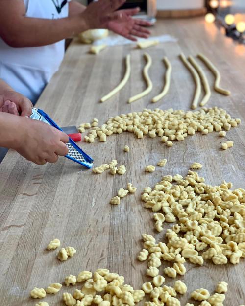 Hands shaping pasta on a wooden surface, with various pasta shapes scattered around.