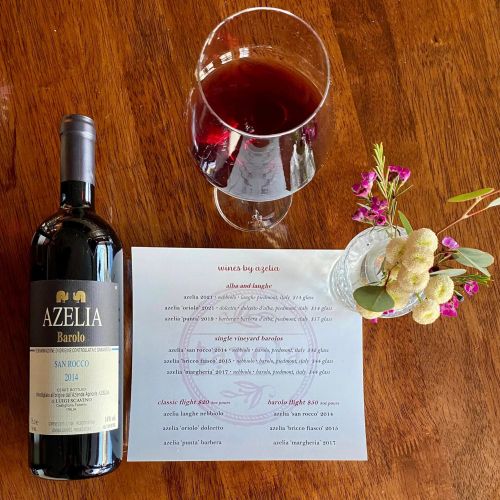 A bottle of Azelia Barolo wine, a glass of red wine, and a small vase with flowers on a wooden table.