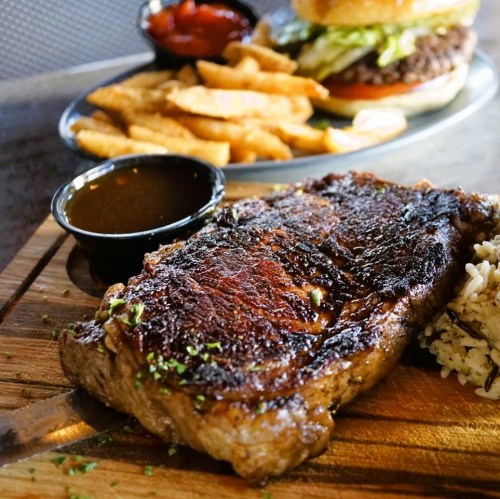 A juicy steak on a wooden board, served with rice and a side of sauce, with fries and a burger in the background.