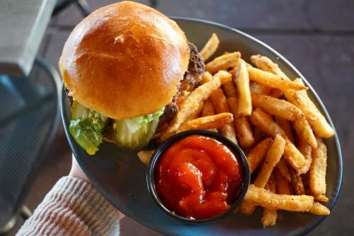 A hand holding a plate with a burger topped with lettuce and pickles, alongside a serving of fries and ketchup.