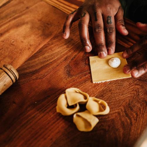 A pair of hands with tattoos shapes pasta dough on a wooden surface, with a small ball of filling nearby.