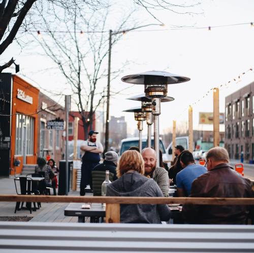 Outdoor dining scene with people at tables, heaters, and a restaurant in the background during sunset.
