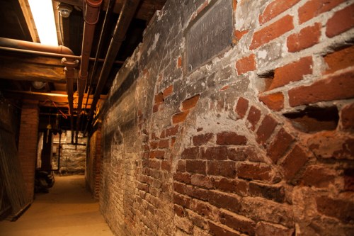 A dimly lit brick basement corridor with exposed pipes and a weathered wall.
