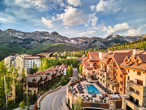 Scenic view of a mountain resort with buildings, a pool, and lush greenery under a blue sky with clouds.