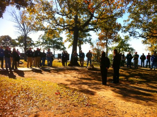 A group of people gathered outdoors under trees, listening to two speakers in a scenic setting.