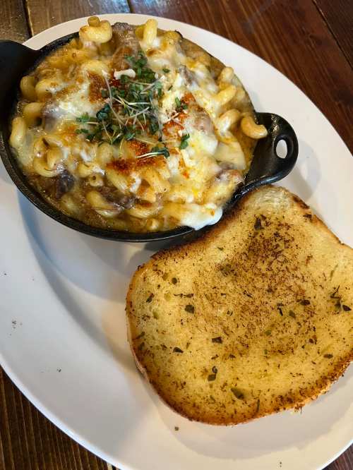 A cast iron dish of cheesy macaroni and beef, topped with herbs, served alongside a slice of toasted garlic bread.