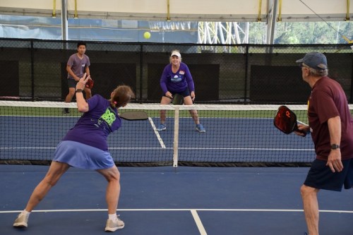 Two women and a man play pickleball indoors, with one woman preparing to hit the ball over the net.