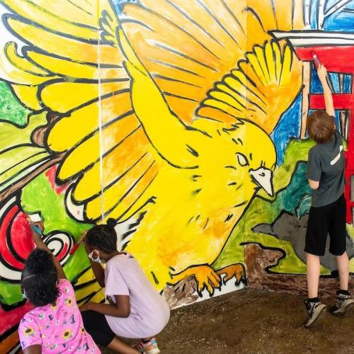 Children painting a vibrant mural featuring a large yellow bird and colorful designs on a wall.