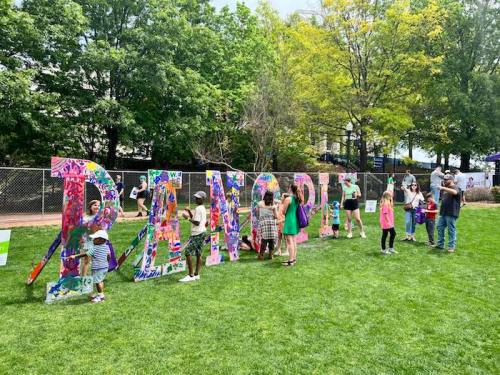 Children and adults gather around a colorful "DANCE" sign in a park, enjoying a sunny day of creativity and fun.