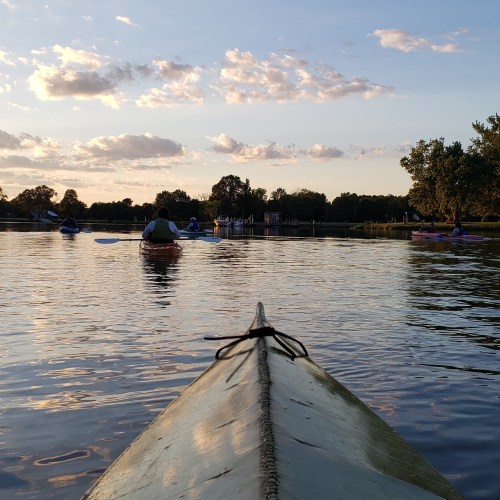 A view from a kayak on calm water, with other kayakers in the distance under a colorful sunset sky.