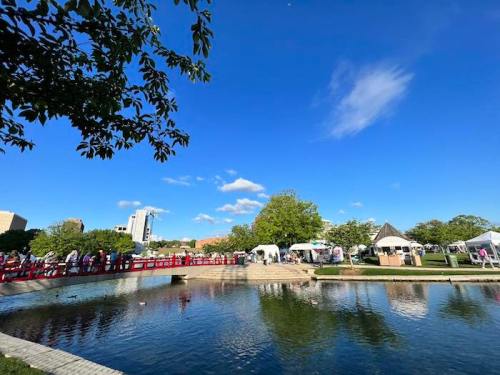 A serene park scene with a pond, red bridge, tents, and blue sky dotted with clouds.