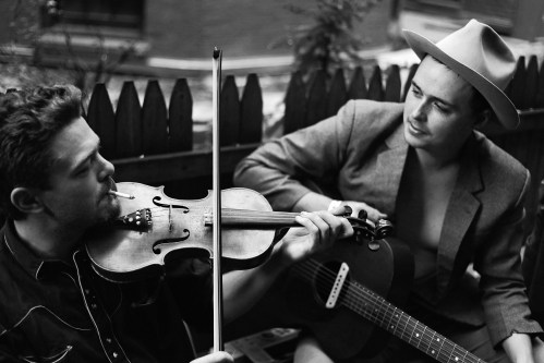 Two musicians play instruments outdoors: one with a violin and the other with a guitar, both in casual attire.
