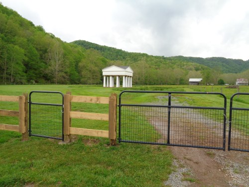 A gated entrance leads to a grassy area with a classical building and green hills in the background.