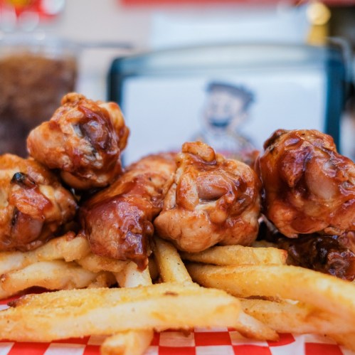A close-up of crispy fried chicken wings glazed in sauce, served with golden French fries on a red checkered paper.