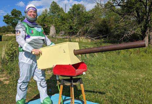 A person in a space-themed costume stands beside a large, homemade cannon in a grassy field.