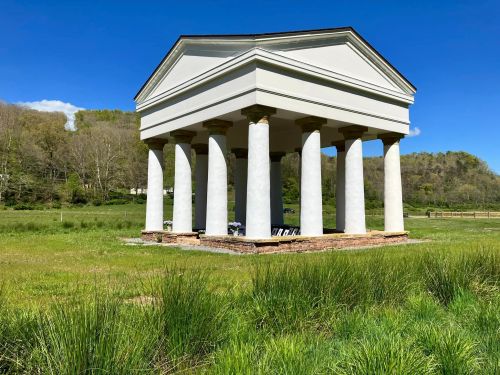 A white, classical-style structure with columns stands in a grassy field under a clear blue sky.