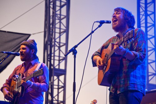 Two musicians perform on stage, singing into microphones while playing acoustic guitars, with a colorful backdrop.